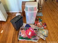 Rubbermaid container filled with holiday wrapping paper rolls stacked vertically, gift bags, tissue paper and assorted wrapping items nearby. Black PS45 shredder visible beside container.