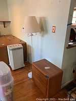 White metal floor lamp with white fabric lampshade next to wooden nightstand and a white appliance.