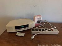 Front view of Bose Wave Music System with digital clock display, remote control in front, power strip and battery tester box behind.