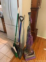 Photo showing Dyson vacuum, Libman broom, and steel core snow shovel leaning against a wall near a refrigerator and grandfather clock on hardwood and tiled floor.