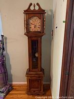 Front view of grandmother clock showing entire height, wooden hood with finial, clock face with numerals, and rectangular base cabinet.