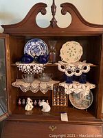 Display cabinet showing various porcelain figurines, blue cobalt glass creamer and sugar, clear glass items, a plate with green clover design, and thimble collection box.