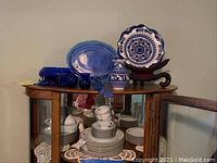 Wide view showing Noritake Joanne China stacked inside glass display cabinet and blue glass bud vases, platters and ceramic pieces on wooden top shelf