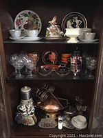 Three shelves with plates, glassware, decorative porcelain flower piece, and kitchen items including a copper wok.