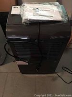 Generations black portable dehumidifier with power cord wrapped around the middle and owner's manual resting on top in plastic wrap.