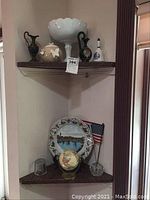 Two corner shelves with assortment of decorative items such as milk glass compote, porcelain bell, decorative ewers, plates, and globe with flag.