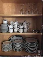 Wood cabinet shelf holding stacks of white plates, bowls, and cups with silver rims, plus two small silver-colored egg cups.