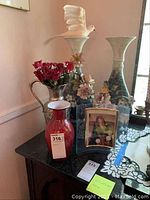 Shows arrangement of 4 vases including 2 ceramic with flower decorations, 1 red glass, 1 metal holding artificial flowers, and framed religious picture on black surface.
