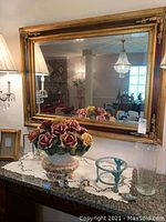 Wide view of large wall mirror hanging above a table with decorative items. Mirror is rectangular with thick ornate gold frame and beveled glass.