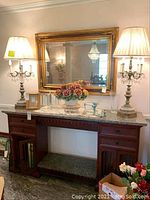 Wide angle showing large rectangular gold-framed mirror mounted on wall above wooden marble-topped console table with drawers, with porcelain flowers and photo frames on table, flanked by two large crystal-lined lamps with white fabric shades