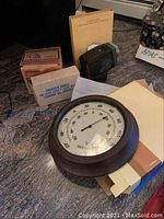 Round Crate & Barrel thermometer with wooden frame resting on some cardboard and paper on floor, thermometer face shows temperature in Fahrenheit and Celsius.
