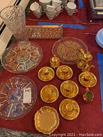 Overview of gold tone tea cups, saucers, creamer, sugar pot, glass cake plates, and vase on a red cloth.