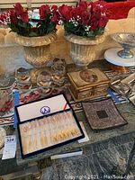 Photo showing two gold-toned urn-style vases holding artificial red roses and a collection of six gold leaf decorated drinking glasses, decorative jewelry box, and boxed Baek San cutlery set on a table.