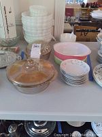 Overall view of sale items on table showing milk glass stack, Pyrex baking dishes in flamingo pink and lime green, clear lid casserole and floral plates stack