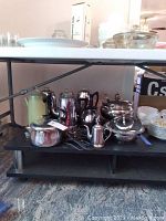 Full shot showing 6 metal coffee pots, including electric coffee makers, a pastel green Faberware kettle, a creamer, a covered serving dish, and a stack of small plates under a table.