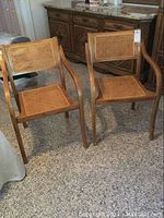 Pair of vintage wooden armchairs with caned seats and backs, shown side by side on carpeted floor near wooden dresser.