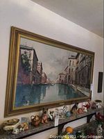 Long image showing multiple animal figurines, paperweight, assorted on shelf beneath a large painting of a canal scene.