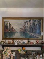 Framed signed print of Venice canal scene hanging on wall above a wooden shelf with various decorative items.