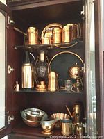 Front view of cabinet shelf with copper-clad tea and coffee service items including teapots, mugs, seltzer bottle and glass carafe with copper frame, silver-plated bowl, and other serving pieces.