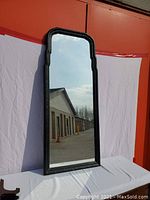 Full front view of tall mirror standing on a white cloth-covered surface against an outdoor backdrop. Shows its arched top and scalloped black wooden frame with floral carvings.