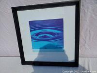 Framed artwork showing blue waterdrop ripple image, black frame with white matting, photographed outdoors with shadow visible.