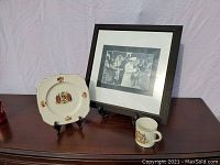 Full view of the lot showing commemorative plate on stand, framed photo of Queen's visit to Canada on stand, and commemorative cup, placed on wooden surface with neutral backdrop.