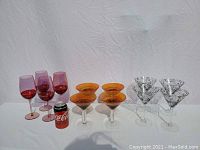 Photo showing all twelve glasses arranged in three groups: four red wine glasses, four orange martini glasses, and four clear martini glasses with floral pattern, alongside a soda can for scale.