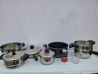 Full view of 9 pots with lids, measuring cup and small containers, showing variety of sizes and styles including stainless steel and black non-stick material.