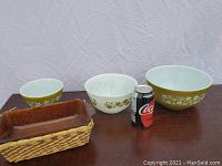 Three Pyrex bowls of various sizes with Spring Flowers pattern and amber bread pan with woven basket alongside a soda can for scale.