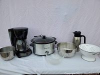 The full lot showing the Bravetti crock pot, Mr. Coffee coffee maker, Nissan thermos, stainless steel mixing bowls, clear glass mixing bowl, and white metal colander against a white backdrop.