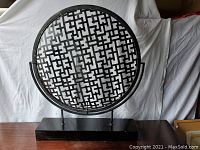 Front view of large round decorative metal screen with geometric openwork design, mounted on rectangular black wood base with small chip in paint.