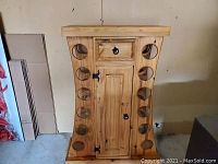 Front angle view of rustic pine wine bar showing eight circular bottle slots and cabinet door with drawer above, iron hardware and red wine stain on base.