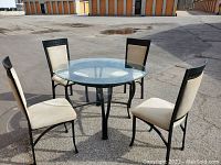 Full view of round glass dining table with four beige upholstered wrought iron chairs around it, set outdoors on concrete.