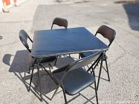 Photo showing the black folding table and four matching chairs positioned outdoors with clear view of table top and chair frames.