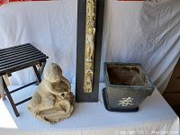 Photo showing small black wooden slatted plant stand, concrete garden centerpiece statue of child and dog, vertical carved stone wall hanging, and square planter pot with white Asian character.