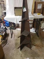 Tall dark stained wooden folky whatnot shelf shown full view hanging from a stand, highlighting its triangular shelves and peg supports.