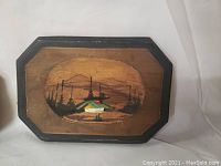 Wooden wall plaque with a painted cabin under trees and mountains in warm tones, black bordered octagonal shape.