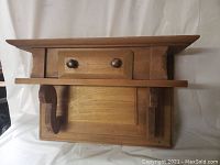 Front and top view of a butternut wood wall shelf with a small central drawer and side supports, shown against a fabric backdrop.
