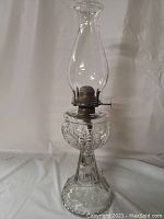 Full view of clear glass oil lamp with metal burner and chimney on white background.