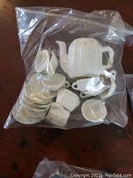 Plastic bag containing white miniature wheat pattern doll dishes, including teapot, saucers, cups, trays