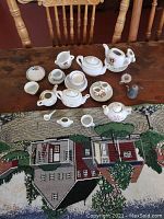 Top view of the 25 piece doll dish set arranged on a wooden table and partially on a woven tapestry with a house design. Items include multiple teapots, sugar bowls, creamer, cups, saucers, small plates, and spoons.
