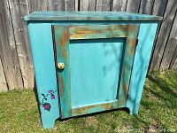 Front view of antique painted pine cupboard showing turquoise paint, distressed finish, single door with round knob, and floral design painted vertically on the left side.