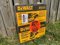 Shrink-wrapped set of two DeWALT sander boxes stacked side by side outdoors against wooden fence.