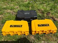 Three hardshell cases (two yellow DeWalt and one black Bostitch) closed and placed on grass.