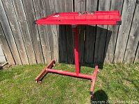 Red metal mechanics parts table standing outdoors on grass near wooden fence, showing structure and tray surface.