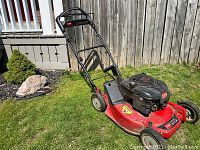 Side angle view of the full lawn mower showing red deck and black engine cover with handlebars and grass catcher.