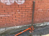 Full view of the orange heavy duty car jack showing the vertical steel bar with holes and handle lying horizontally on pavement in front of a brick wall.