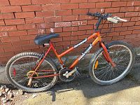 Full side view of an orange Supercycle SC1700 mountain bike leaning against a brick wall, showing frame, tires, pedals, and seat clearly.