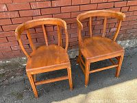 Pair of old wooden chairs with curved backs leaning against red brick wall showing front angle and wear.