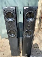 Pair of black Quest Q665 floor standing speakers with three visible drivers each, shown from front in daylight with slight cabinet edge wear visible.